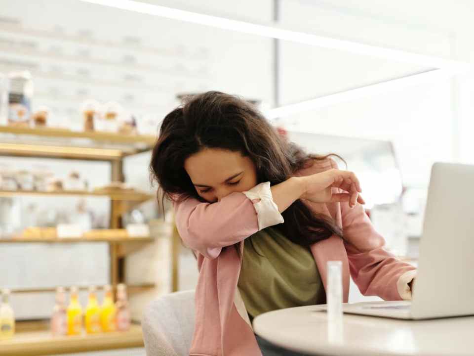 Mulher em mesa de escritório tampando a tosse com o braço.