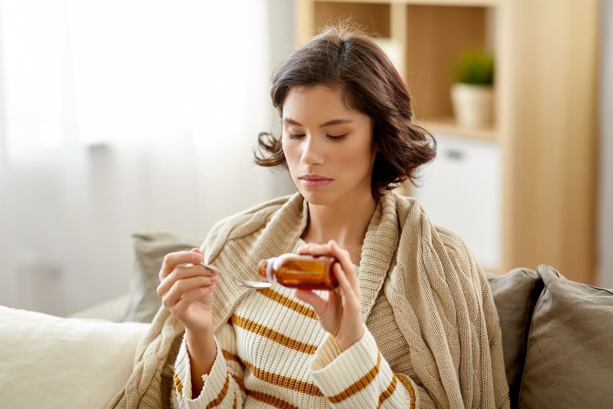 Mulher sentada no sofá doente tomando xarope para tosse.